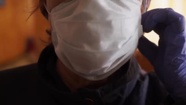 An elderly woman puts off a protective medical mask near the window, looks at the camera. COVID-19 pandemic coronavirus prevention. Social distancing - Filmagem, Vídeo