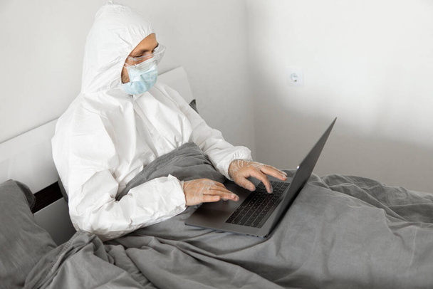 Man in protective white suit and medical mask is working from home in a bed with laptop because of coronavirus epidemic. Remote work during pandemic. Stay home during COVID-19 quarantine concept. - Fotó, kép