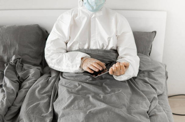 Man in protective white suit and medical mask is working from home in a bed with a tablet because of coronavirus epidemic. Remote work during pandemic. Stay home during COVID-19 quarantine concept. - Foto, immagini