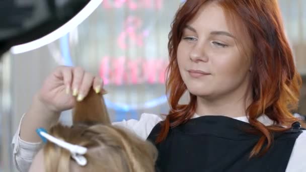 Prachtige roodharige meester werken met het haar van een klant doet een krullend kapsel met behulp van een krultang. De kunstenaar creëert een krullen kapsel voor lang blond haar in een beauty studio. - Video
