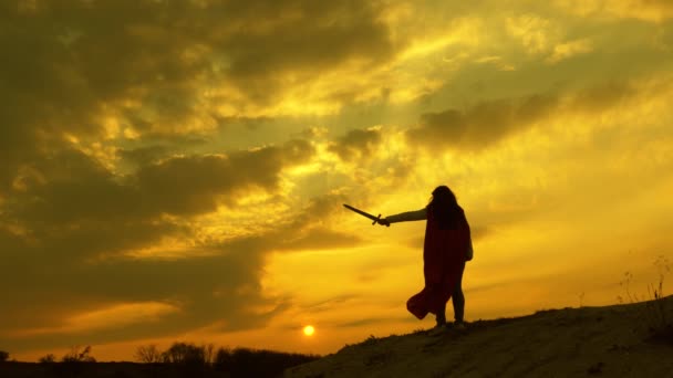 svobodná žena s mečem v ruce a v červeném plášti stojí na hoře a dívá se na západ slunce. volná dívka hraje superhrdinu. dívka hraje romlenina v červené paprsky slunce proti obloze - Záběry, video