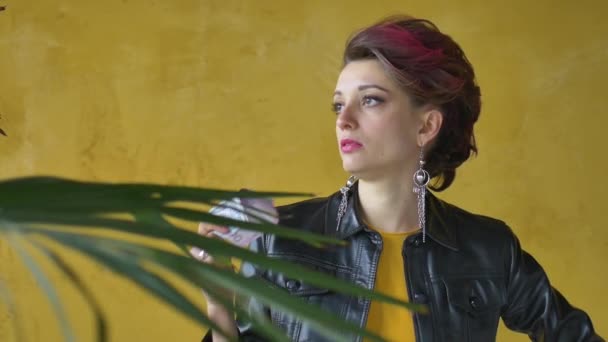 Glamouröse aggressive Dame in Punk-Rock-Party-Kleidung mit dunkelrosa Haaren in schwarzer Lederjacke und langen Ohrringen auf gelbem Hintergrund mit Geld-Dollars und Griwna in den Händen - Filmmaterial, Video