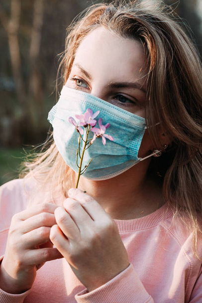 A young blonde woman inhales the smell of a flower through a medical mask. - Photo, Image