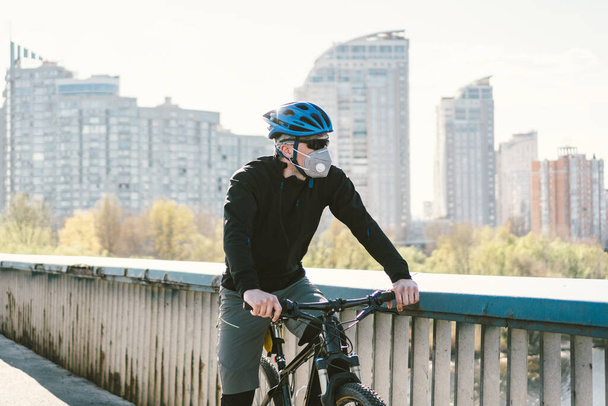 Samiec rowerzysta na rowerze górskim w masce ochronnej respiratora w zanieczyszczonym mieście. Facet jedzie do pracy nad przyjaznym dla środowiska transportem, miejską eko rafą. Maska zanieczyszczająca twarz smog dust pm2.5 ochrona. - Zdjęcie, obraz