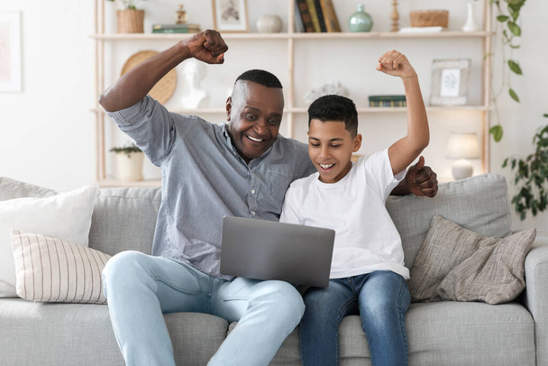 Ώριμος μαύρος μπαμπάς και μικρός γιος βλέποντας αθλητικά στο laptop και επευφημίες - Φωτογραφία, εικόνα