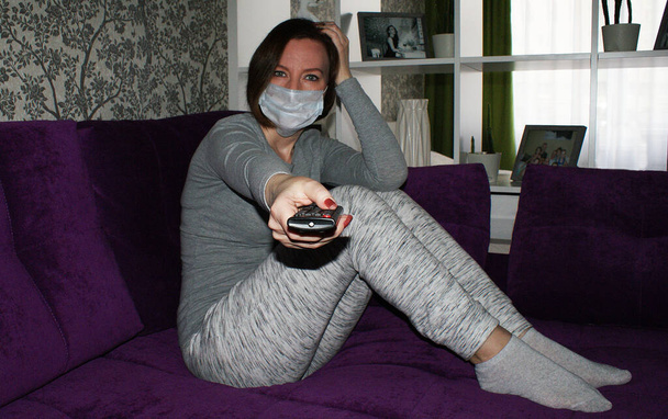 Dívka v karanténě v lékařské masce doma sedí na gauči a dívá se na televizi - Fotografie, Obrázek
