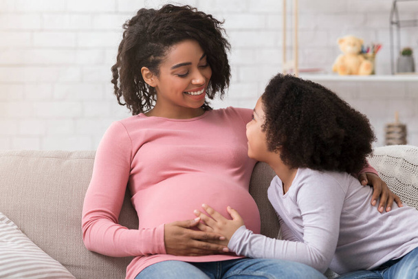 Δεύτερη εγκυμοσύνη. Έγκυος μαύρη γυναίκα που δένεται με τη μικρή κόρη στο σπίτι - Φωτογραφία, εικόνα