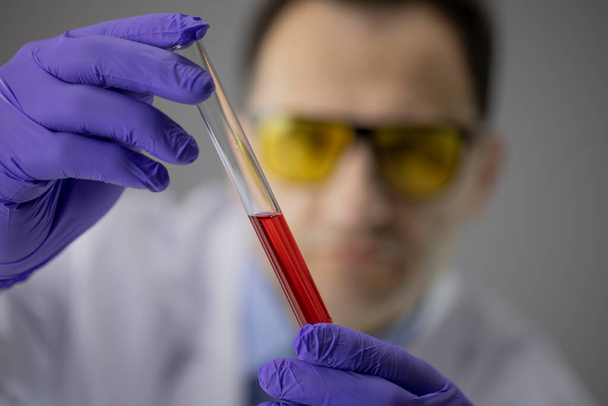 Дослідник хімії в синіх рукавичках, що тримає пробірку з прозорою червоною рідиною
 - Фото, зображення