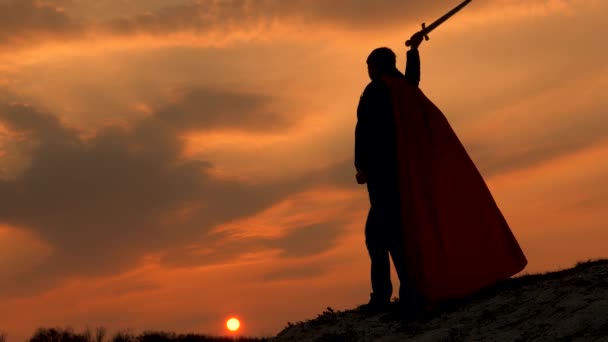 Kırmızı pelerinli bir savaşçı bir dağda duruyor ve gün batımında kılıcını sallıyor. Özgür erkek şövalye. Özgür adam süper kahramanı oynuyor. Roma lejyonunun parlak güneş ışınlarıyla gökyüzüne karşı oyunu - Video, Çekim