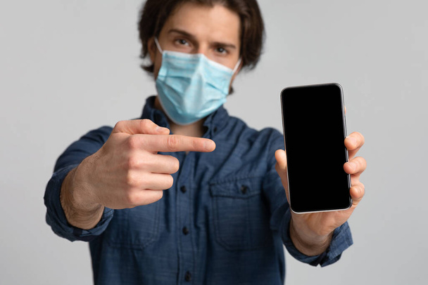 Hombre joven en la máscara quirúrgica que apunta a Smartphone con pantalla negra
 - Foto, Imagen