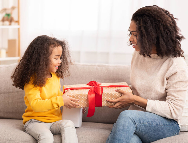 African Mother Giving Gift To Daughter Sitting On Sofa Indoor - Photo, Image