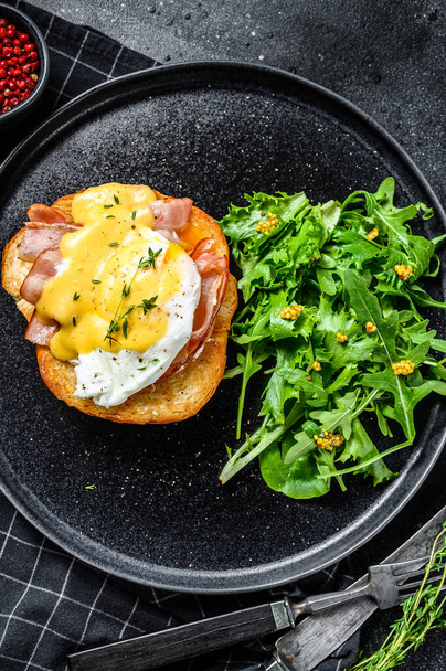 Brioche sandwich with bacon, egg Benedict and hollandaise sauce. Garnish with arugula salad. Black background. Top view. - Photo, image