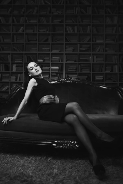 A beautiful brunette girl, a model in the studio. sun and shadow. Standing in a sexy dress with a red banter. Sits on a luxurious couch in black tights. the photo is black white. - Фото, зображення