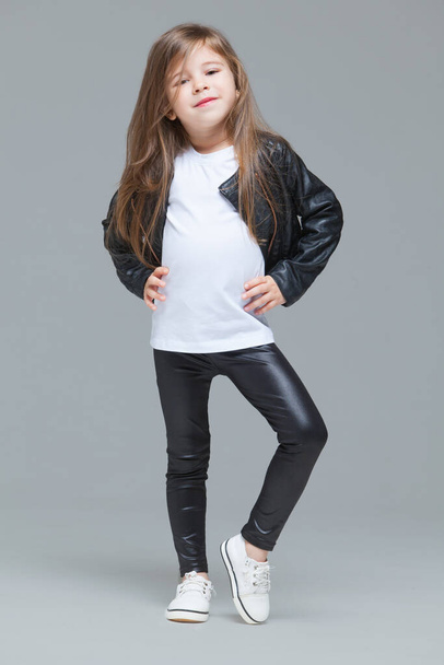 Baby girl with long hair in black leather jacket and leggings is standing in the studio on grey background - Photo, Image