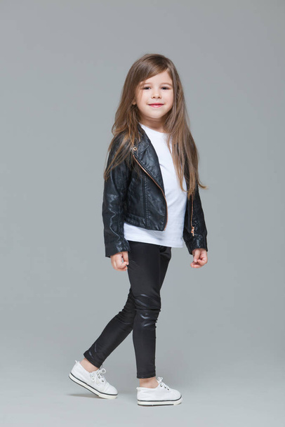 Baby girl with long hair in black leather jacket and leggings is standing in the studio on grey background - Zdjęcie, obraz