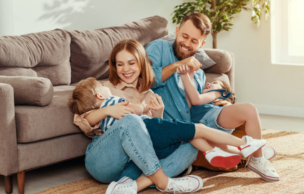 Feliz padre y madre con niños pequeños sentados en el suelo cerca del sofá y riendo mientras se divierten juntos en casa durante weeken
 - Foto, Imagen