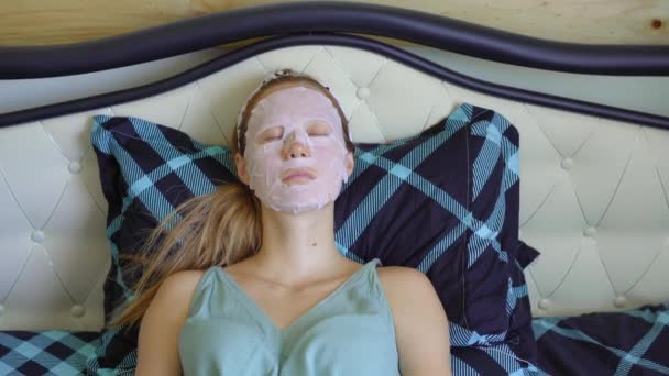 A young woman laying on a bed with a cosmetical face mask on her face. Skincare concept - Záběry, video