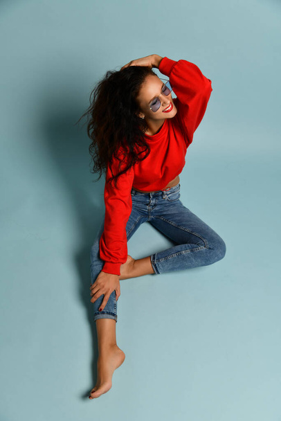 Happy adult woman with curly brunette hair in smoked blue glasses, red sweater and blue jeans is sitting on the floor - 写真・画像