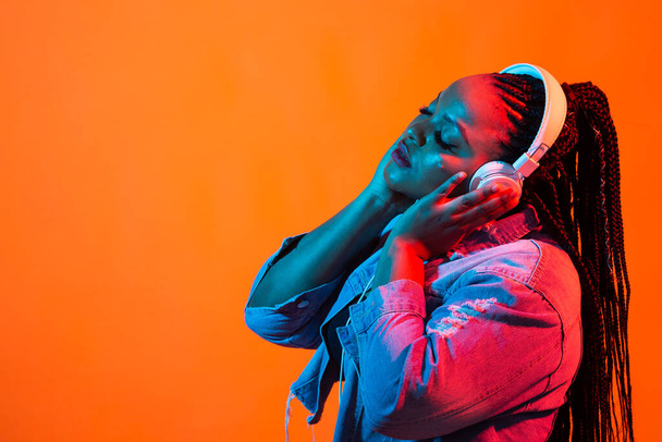 Ritratto al neon di una giovane donna africana che ascolta musica con gli auricolari. Sfondo arancione con spazio di copia
. - Foto, immagini