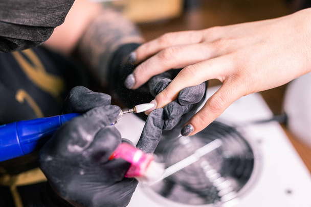 Zbliżenie manikiurzystki usuwa żelowy lakier szelakowy z paznokci klientów za pomocą manikiurzystki. Manicure master pracuje z elektrycznym wiertłem do paznokci w salonie piękności. Proces manicure sprzętowy. - Zdjęcie, obraz