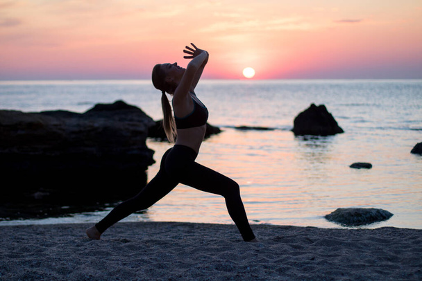 girl has a yoga practice on the beach among big stones. rocks in the sea. sunrise time - Photo, Image
