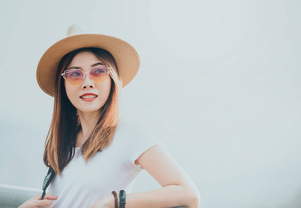 Молодая азиатская женщина в шляпе и солнцезащитных очках, глядя в сторону с счастливой улыбкой в сторону пустого белого копировального пространства, путешествия на открытом воздухе концепции
. - Фото, изображение