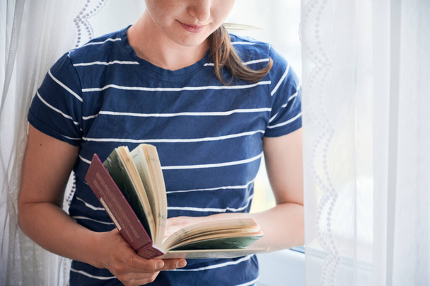 Cropped close-up snapshot of young girl standing next to the window reading book in home isolation during covid-19 crisis. Self education in pandemic 2020. Stay at home save lives - Photo, image
