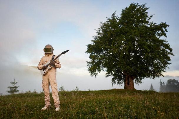 Full length of cosmonaut guitarist with musical instrument standing in hillside meadow with beautiful tree and sky on background. Astronaut in space suit and helmet playing guitar outdoors. - Фото, изображение