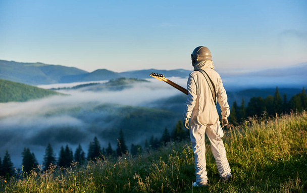 ギターを持った宇宙旅行者のバックビューは、芝生の丘に立って、美しい霧の山の景色を楽しんでいます。男性宇宙飛行士ギタリストで宇宙服姿で霧の多い丘を青空の下で見る. - 写真・画像
