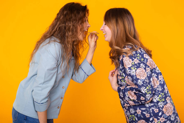 Two funny female friends or sisters eating sharing cookie on yellow background. Friendship, relationship and fun concept. - Foto, Imagem