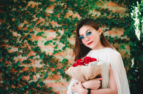 Usmívající se dívka s kyticí rudých růží, Portrét spokojené mladé ženy oblečené v bílých šatech držící kytici růží - Fotografie, Obrázek