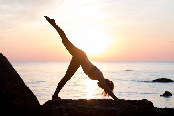 Mädchen hat eine Yoga-Praxis am Strand zwischen großen Steinen. Felsen im Meer. Sonnenaufgangszeit - Foto, Bild