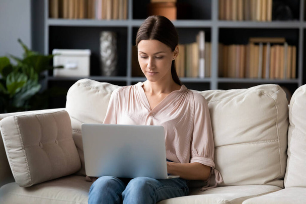 Serious young woman using laptop, sitting on couch at home - Foto, Imagen
