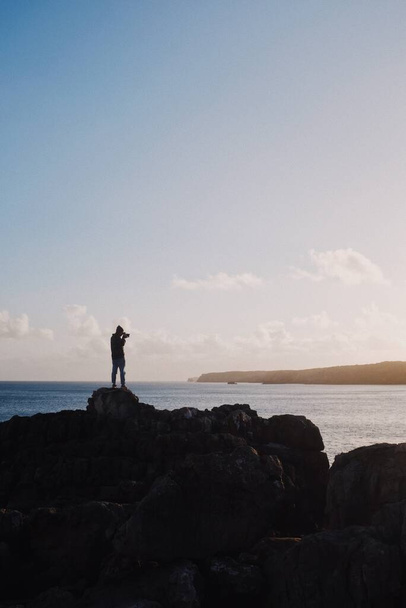 A vertical shot of a person standing on a rock at the ocean and taking a picture - Foto, Imagem