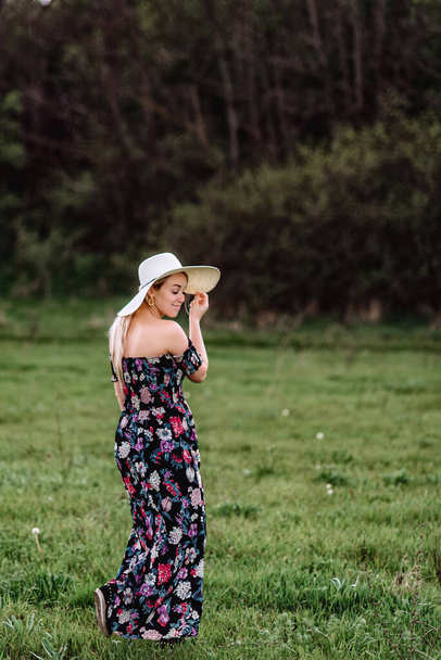 a beautiful girl in a dress and a hat walks alone in a green meadow. Young pretty woman blonde outdoors enjoy freedom and relaxation - Foto, afbeelding