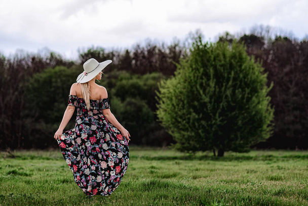 uma menina bonita em um vestido e um chapéu caminha sozinha em um prado verde. Jovem mulher bonita loira ao ar livre desfrutar de liberdade e relaxamento
 - Foto, Imagem