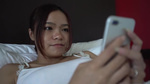 4K Asian woman lying on the bed with white sheets and using smartphone in a beautiful hotel room. Surfing the internet, checks social network and see pictures on her phone device. -Dan - Filmati, video
