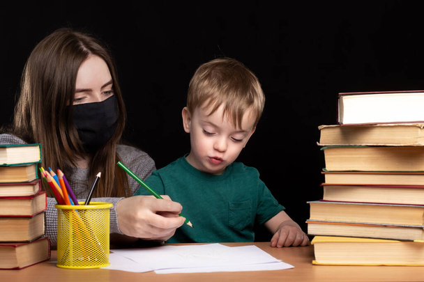 Mam met een medisch masker helpt haar zoon te leren schrijven. aan een tafel met boeken op een zwarte achtergrond. Afstandsonderwijs in quarantaine. moeilijkheden met online lessen. - Foto, afbeelding