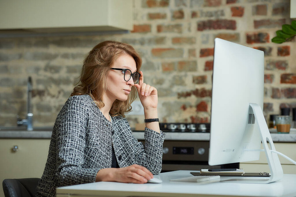 Une femme en lunettes travaille à distance sur un ordinateur de bureau dans son studio. Une femme patronne à la recherche d'informations lors d'une vidéoconférence à la maison. Une jolie professeure se prépare pour une conférence en ligne
. - Photo, image