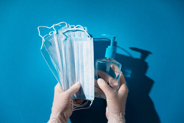 Woman hands in latex gloves holding facial medical masks and sanitizer, blue background - Fotografie, Obrázek