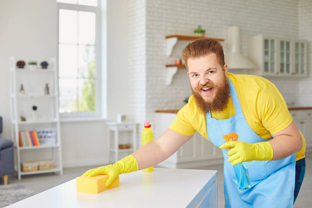 Funny fat man in an apron and yellow cleaning gloves is cleaning in the room. - Photo, image