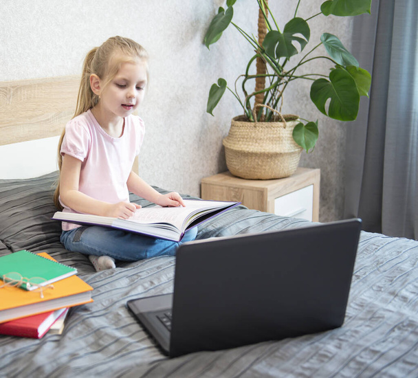 Biała dziewczyna dziecko siedzi w łóżku i uczy się online na laptopie. Szkoła domowa, edukacja online, edukacja domowa, kwarantanna, koncepcja koronawirusa. - Zdjęcie, obraz