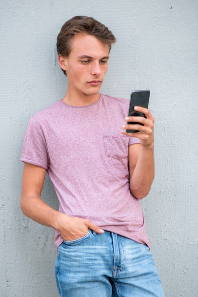 Портрет серйозний молодий чоловік спирається на стіну, дивлячись на мобільний телефон
 - Фото, зображення