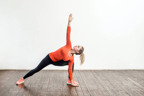 Slim blond woman in tight sportswear practicing yoga, standing in trikonasana triangle pose, training muscles for flexibility. Health care, sports activity and workout at home. indoor studio shot - 写真・画像