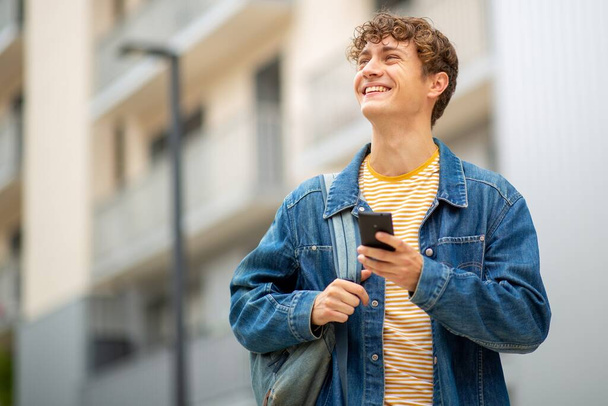 Portret lachende jongeman loopt met tas en mobiele telefoon buiten - Foto, afbeelding