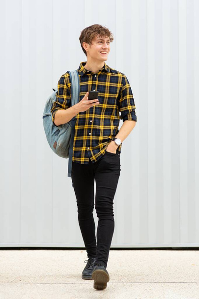 Retrato de cuerpo entero joven feliz caminando con bolsa y teléfono celular por fondo blanco
 - Foto, Imagen