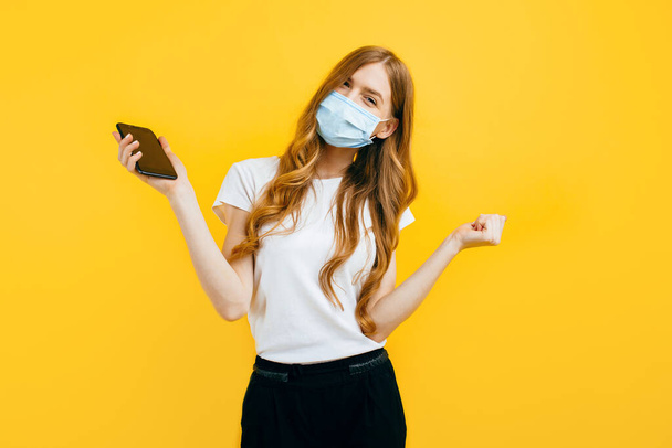 A young woman, wearing a protective medical mask, uses a mobile phone on a yellow background. The concept of quarantine, coronavirus - Photo, Image