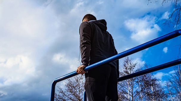 athlete in training. A man in a sweatshirt on the sports ground. silhouette of a man on uneven bars. Street workouts. Sports activities on the street. man on a background of blue sky. - Fotoğraf, Görsel