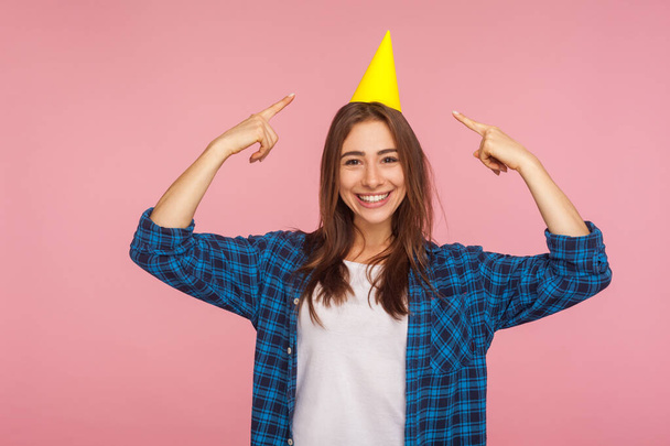 Portrait of cheerful pretty girl pointing to funny cone on head and smiling, celebrating New Year holiday or anniversary, expressing joy, festive mood. indoor studio shot isolated on pink background - Photo, image