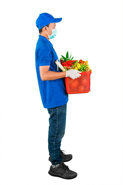Side view of man wearing delivery uniform and face mask while holding a red basket full of fruits & vegetables, isolated in white background - 写真・画像